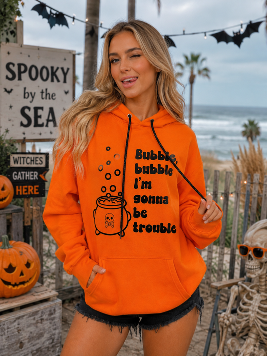 A woman wearing a bright orange hoodie with a black graphic of a cauldron and the text 'Bubble bubble I'm gonna be trouble' stands outdoors at a beach-themed Halloween display with pumpkins, signs, and a skeleton.
