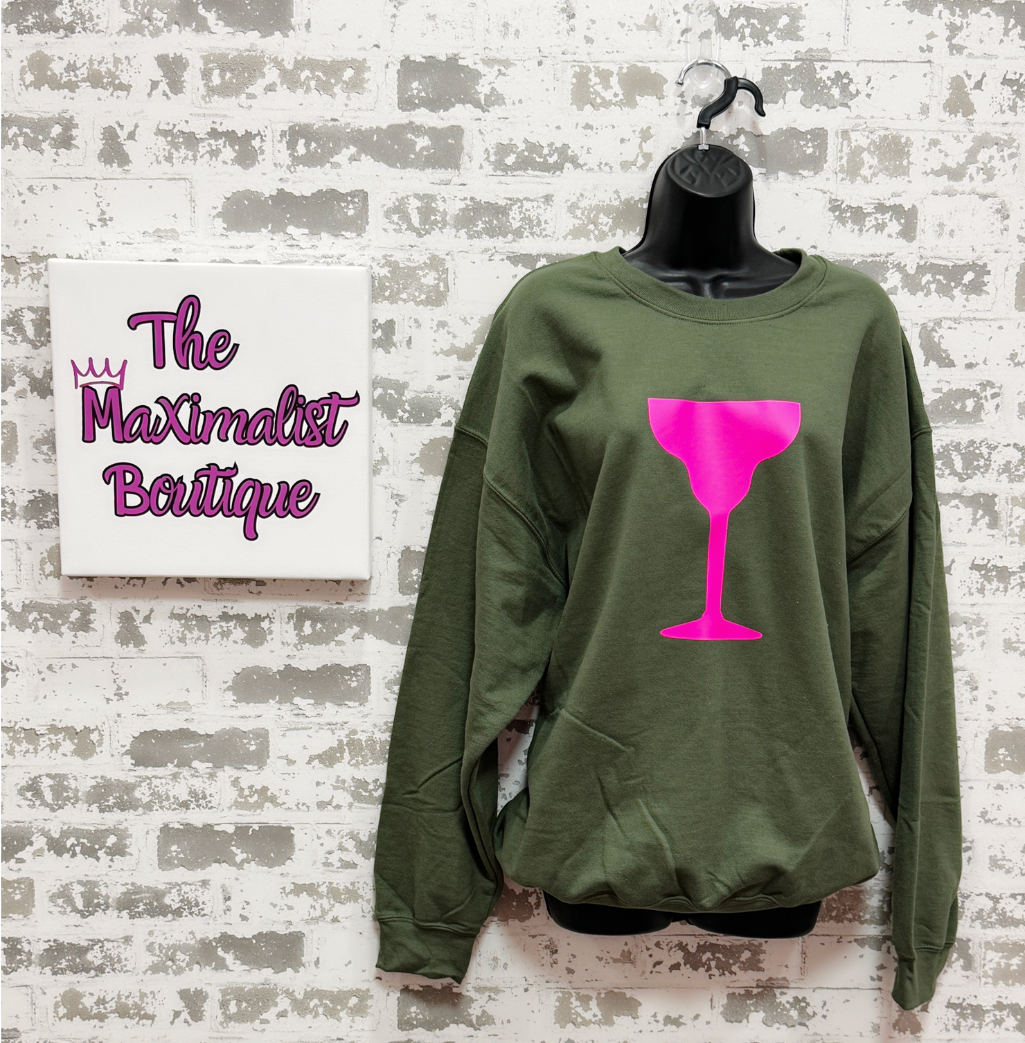 Green sweatshirt with pink wine glass design on a hanger against a textured wall with 'The Maximalist Boutique' sign.