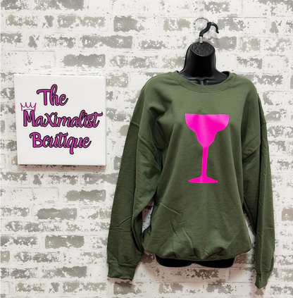 Green sweatshirt with pink wine glass design on a hanger against a textured wall with 'The Maximalist Boutique' sign.