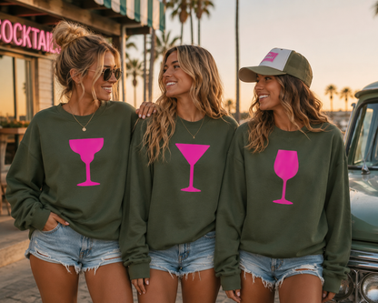Three women standing together outdoors at sunset wearing matching olive green crewneck sweatshirts with large pink wine glass graphics on the front. The woman on the right also wears a green and white baseball cap.