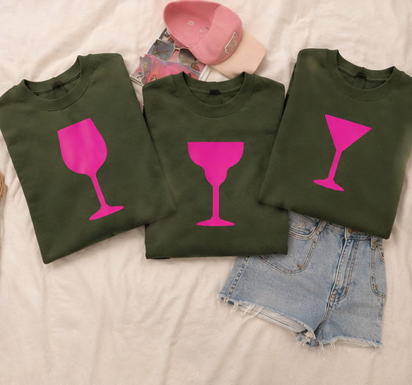 Three olive green crewneck sweatshirts laid flat on a white fabric background, each printed with a bright pink wine glass graphic. A pink cap and denim shorts are styled around them.