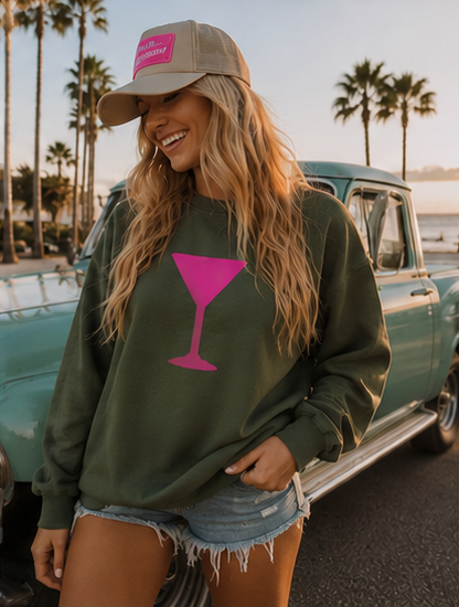 A woman wearing an olive green sweatshirt with a pink martini glass graphic, a khaki baseball cap, and denim shorts, posing in front of a vintage teal pickup truck with palm trees in the background.