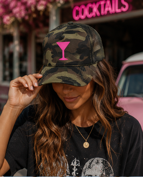 A woman wearing a camouflage trucker hat with a bright pink martini glass graphic on the front, posing outdoors in front of a bar with a pink neon sign.