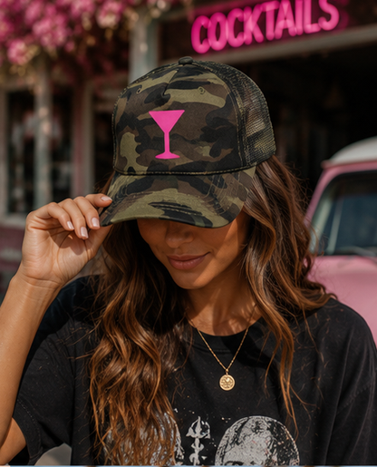 A woman wearing a camouflage trucker hat with a bright pink martini glass graphic on the front, posing outdoors in front of a bar with a pink neon sign.