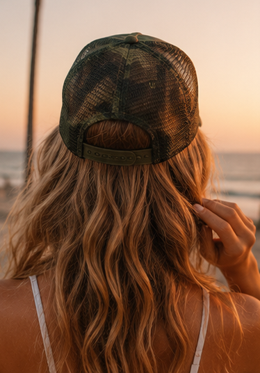 A woman viewed from behind wearing a dark camouflage trucker-style mesh cap, with long wavy blonde hair and a beach sunset in the background.