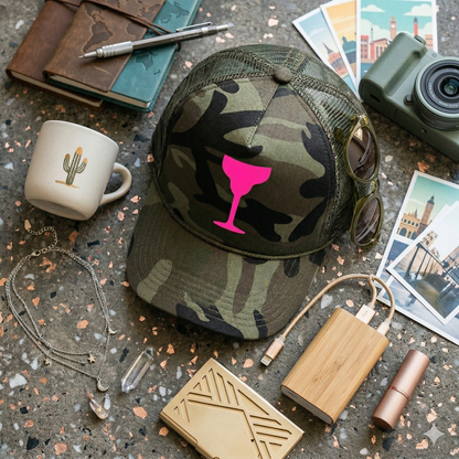 Camouflage cap with pink wine glass design on a textured surface with travel items.