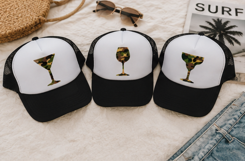 Three black and white trucker hats with camouflage-patterned cocktail glass graphics on the front panels, arranged on a white textured blanket with sunglasses, a woven bag, and denim shorts nearby.