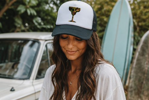 A woman wearing a white and dark gray trucker hat with a graphic of a margarita glass on the front, standing outdoors with a vintage car and surfboards in the background.