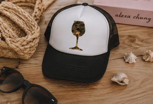 A black and white trucker hat with a wine glass graphic filled with camouflage print, styled on a wooden surface with sunglasses, a woven straw hat, seashells, and a pink box in the background.