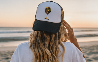 A woman viewed from behind at a beach during sunset, wearing a white and black trucker hat with a graphic of a wine glass filled with camouflage pattern.