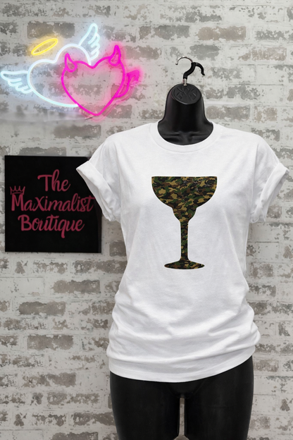 White t-shirt with a camo cocktail glass design on a hanger against a brick wall with neon signs.