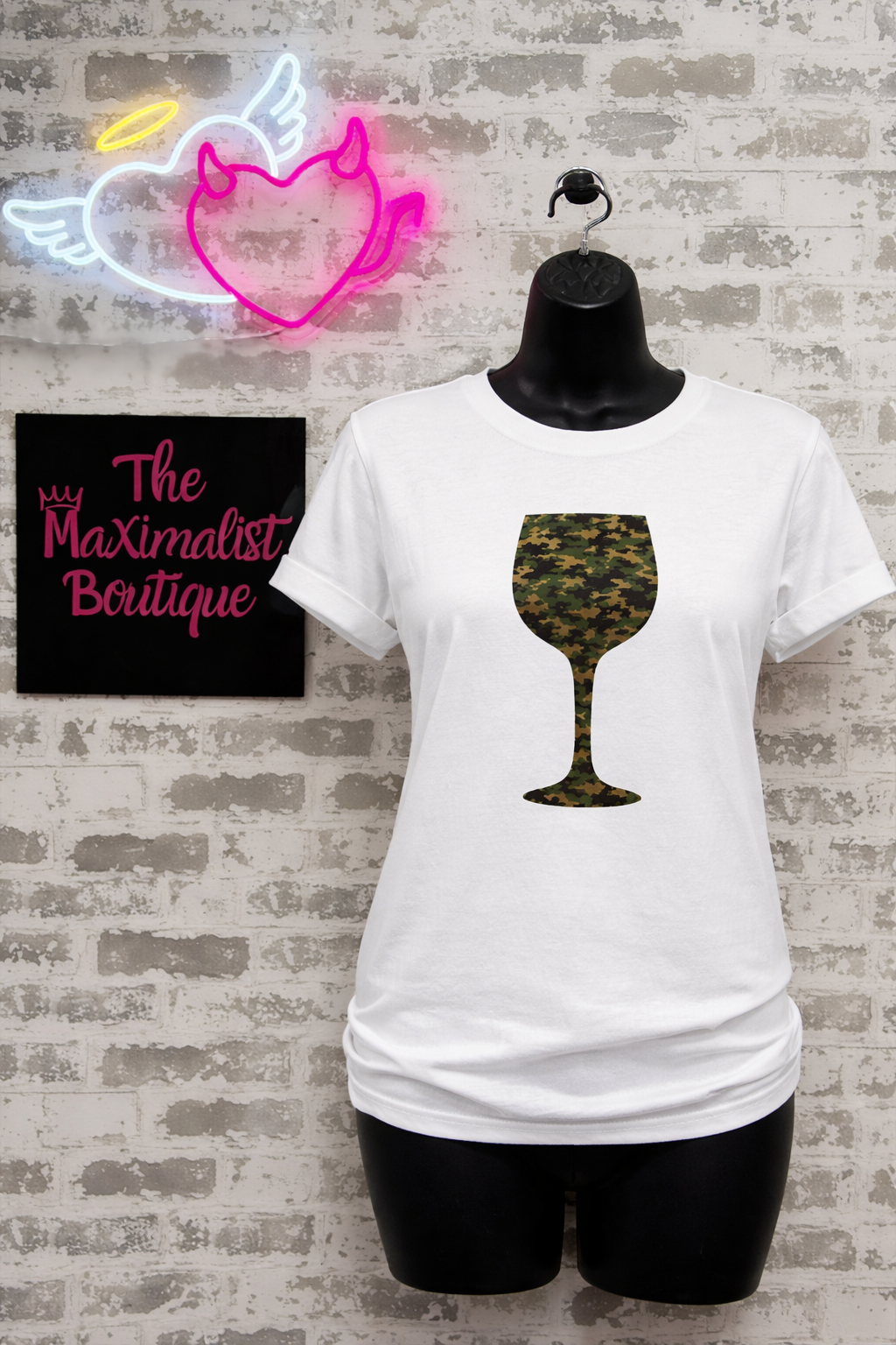 White t-shirt with camouflage wine glass design on a mannequin against a brick wall with neon sign and 'The Maximalist Boutique' sign.