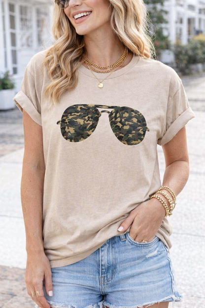 Woman wearing a beige t-shirt with sunglasses graphic and denim shorts outdoors.