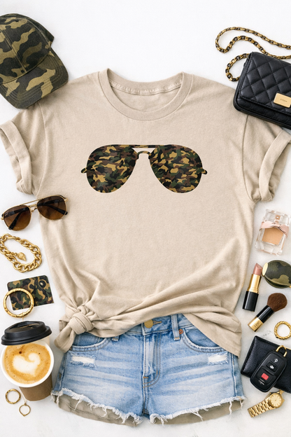 Beige t-shirt with camouflage sunglasses graphic, paired with denim shorts and surrounded by accessories on a white background.