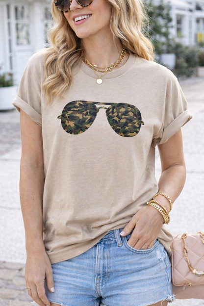 Woman wearing a beige t-shirt with sunglasses graphic and denim shorts, standing outdoors.