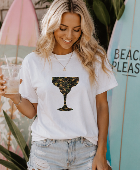 A woman wearing a white graphic t-shirt with a camouflage margarita glass print, paired with light-wash denim shorts, holding a drink in front of surfboards.