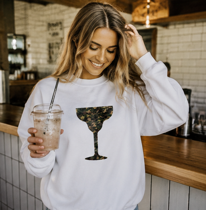 A woman in a white sweatshirt with a camouflage margarita glass graphic, holding an iced coffee in a cafe setting.