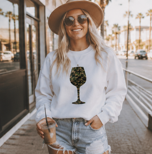 A woman wearing a white crewneck sweatshirt with a green camouflage wine glass graphic, styled with a tan hat, sunglasses, and denim shorts, holding an iced coffee on a sunny city sidewalk.