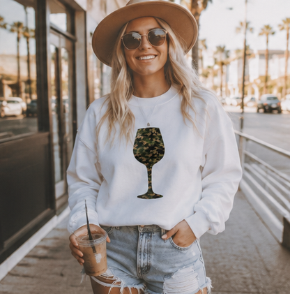 A woman wearing a white crewneck sweatshirt with a green camouflage wine glass graphic, styled with a tan hat, sunglasses, and denim shorts, holding an iced coffee on a sunny city sidewalk.