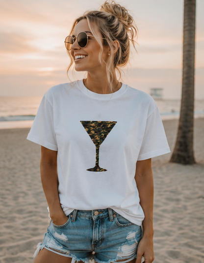 A woman wearing a white graphic t-shirt with a martini glass print in a camouflage pattern, paired with distressed denim shorts, standing on a sandy beach at sunset.