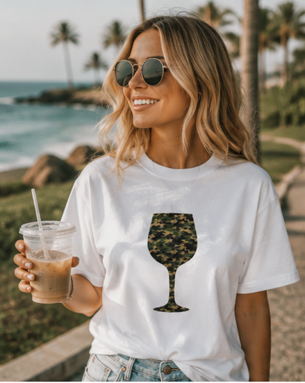 A woman wearing round sunglasses and a white graphic t-shirt with a wine glass design filled with a green camouflage pattern, holding an iced coffee on a beach path with palm trees and the ocean in the background.