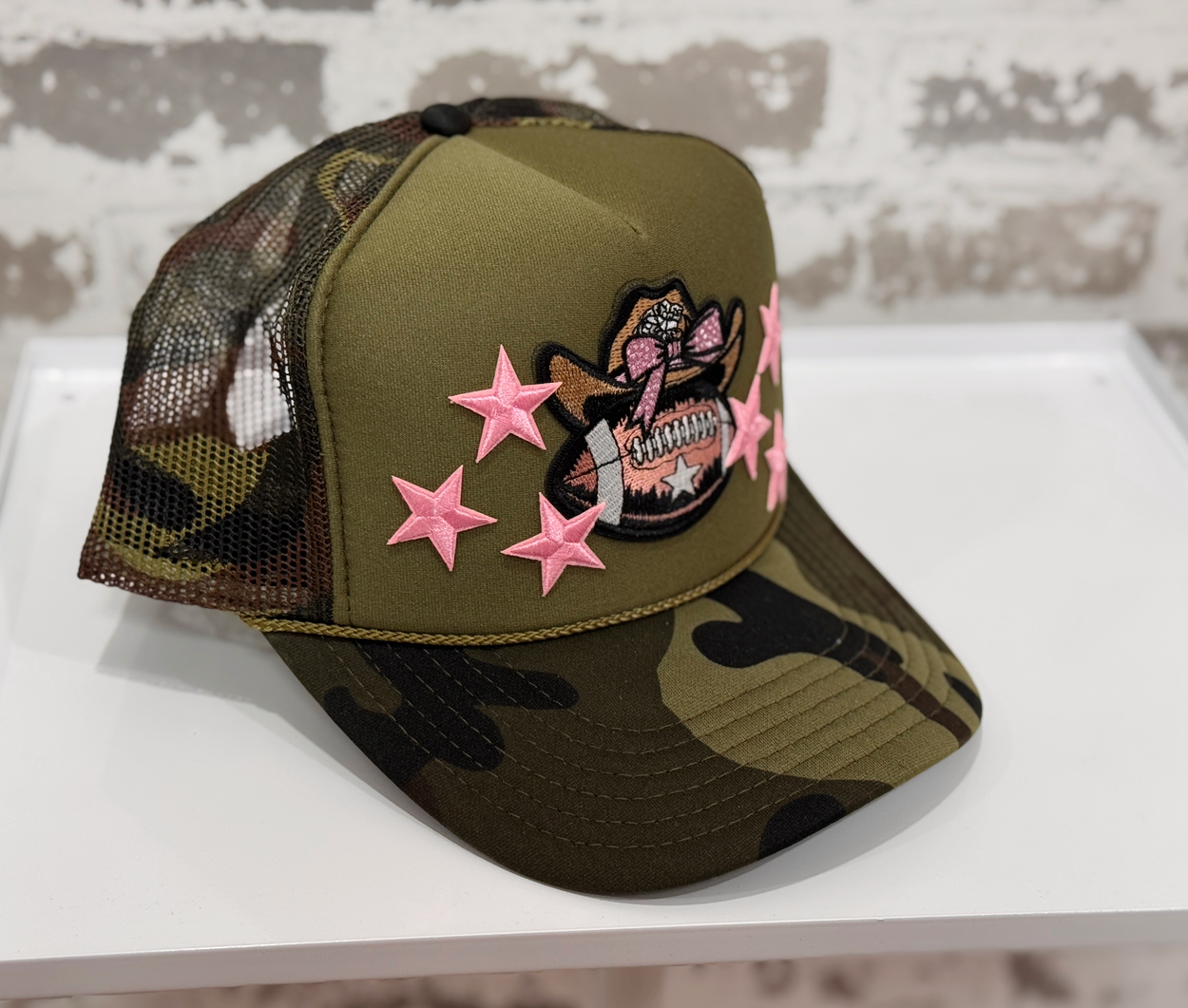 Camouflage cap with pink star designs and a tattoo-like patch on a white surface.