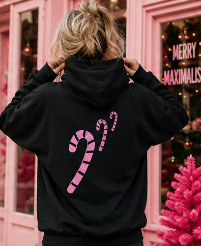 A person viewed from behind wearing a black hoodie with a large pink striped candy cane graphic on the back, standing in front of a pink storefront with holiday decorations.