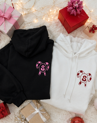 Two folded hoodies, one black and one white, displayed on a white textured blanket with holiday gift boxes, fairy lights, and ornaments. Each hoodie features a candy cane heart graphic with a pink and red letter 'S' in the center.