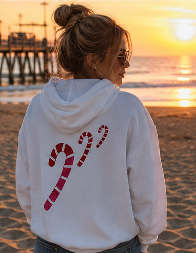 A woman standing on a beach at sunset, viewed from behind, wearing a white hoodie with a large red and white striped candy cane graphic on the back.