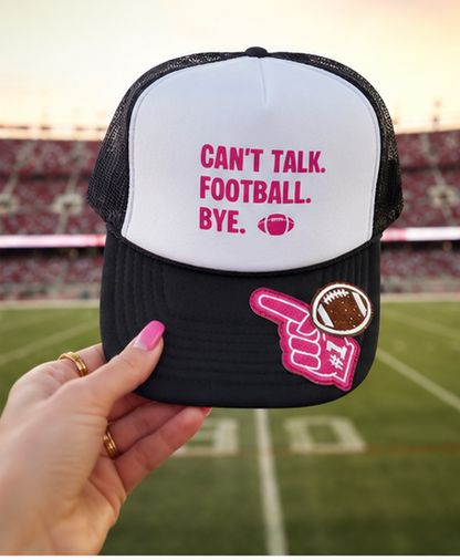 A hand holds a black and white trucker hat with pink text reading 'CAN'T TALK. FOOTBALL. BYE.