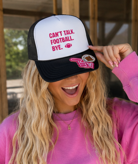 A woman wearing a black and white trucker hat with pink text that reads 'CAN'T TALK. FOOTBALL. BYE.