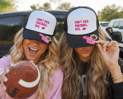 Two women wearing matching trucker hats with the text 'CAN'T TALK FOOTBALL BYE.' in pink on a white front panel and black mesh sides, posed outdoors in front of cars with a football visible in the foreground.