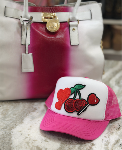 Pink and white cap with cherry design in front of a pink and white handbag.
