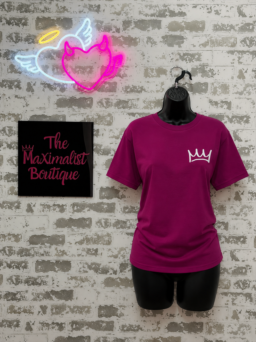 Purple t-shirt with a logo on a mannequin against a textured wall with neon sign and 'The Maximalist Boutique' sign.