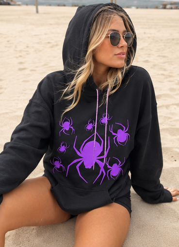 A woman sitting on a sandy beach wearing a black hoodie with a purple spider graphic print and matching sunglasses.