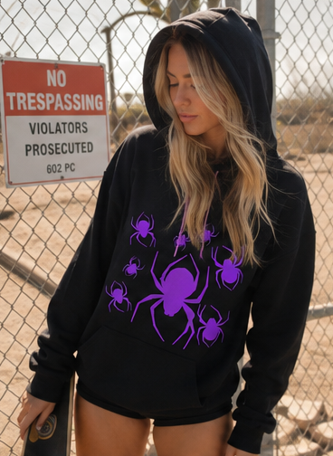 A woman wearing a black hoodie with a purple spider graphic print, standing in front of a chain-link fence and a 'No Trespassing' sign.