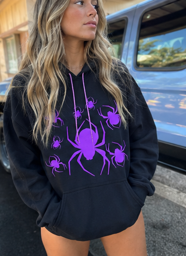 A woman wearing a black pullover hoodie with a purple spider graphic print on the front, standing outdoors next to a blue vehicle.