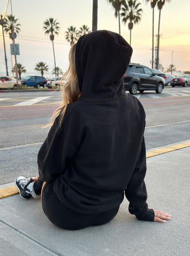 A person sitting on a city sidewalk at sunset, viewed from behind, wearing a black hooded sweatshirt and black pants with black-and-white sneakers. Palm trees, parked cars, and street crosswalks are visible in the background.