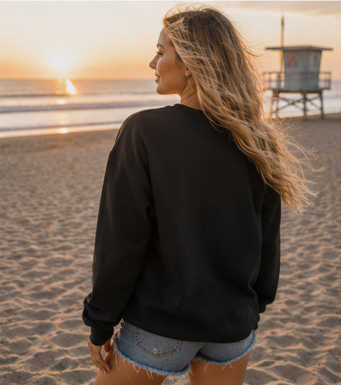 A woman with long blonde hair stands on a sandy beach at sunset, wearing a black crewneck sweatshirt and denim shorts, looking out toward the ocean and a lifeguard tower.