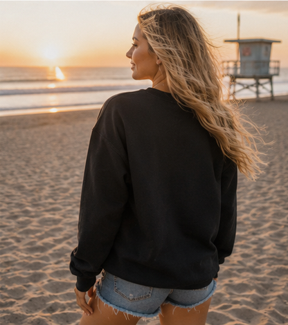 A woman with long blonde hair stands on a sandy beach at sunset, wearing a black crewneck sweatshirt and denim shorts, looking out toward the ocean and a lifeguard tower.