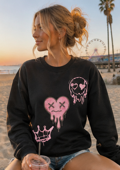 A woman wearing a black crewneck sweatshirt with pink cartoon-style graphics (a heart, skull, and crown) sitting on a beach at sunset.
