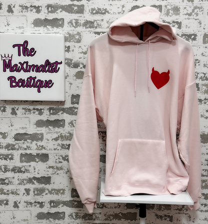 Pink hoodie with a red heart design on a brick wall background with 'The Maximalist Boutique' sign.