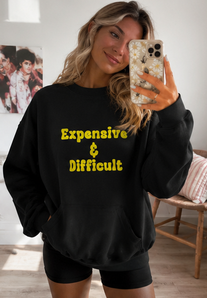 A woman takes a mirror selfie wearing a black sweatshirt with the phrase 'Expensive & Difficult' printed in yellow text on the front, paired with black shorts in a brightly lit room.