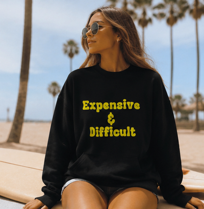 A woman wearing a black crewneck sweatshirt with yellow text reading 'Expensive & Difficult' sits on a beach with palm trees in the background.