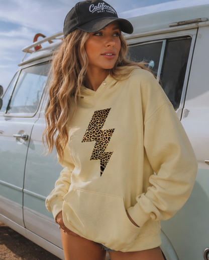 A woman wearing a cream-colored pullover hoodie with a leopard-print lightning bolt graphic on the front, paired with a black baseball cap and standing next to a vintage light blue vehicle.
