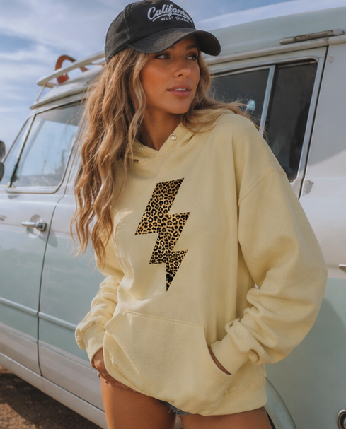 A woman wearing a cream-colored pullover hoodie with a leopard-print lightning bolt graphic on the front, paired with a black baseball cap and standing next to a vintage light blue vehicle.