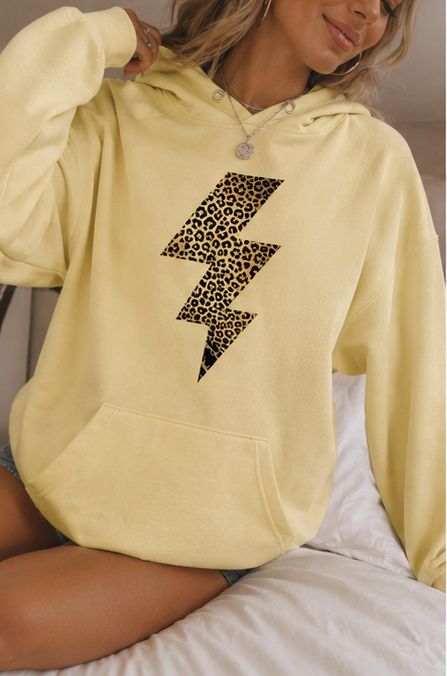 A woman wearing a pale yellow pullover hoodie with a large leopard-print lightning bolt graphic on the front, sitting on a bed with white sheets.
