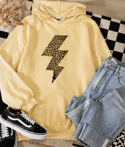 A pale yellow pullover hoodie laid flat with a large black-and-brown leopard-print lightning bolt graphic on the chest. The hoodie is styled with light-wash denim shorts, black-and-white sneakers, and a checkered rug.