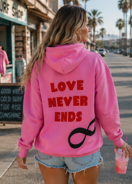 A woman viewed from behind wearing a bright pink hoodie with the words 'LOVE NEVER ENDS' in red and a black infinity symbol on the back, walking on a sunny street with palm trees and storefronts in the background.