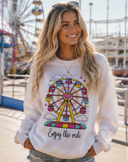 A woman wearing a white crewneck sweatshirt with a colorful graphic of a Ferris wheel and the text 'Enjoy the ride', set against a blurred outdoor background of an amusement park.